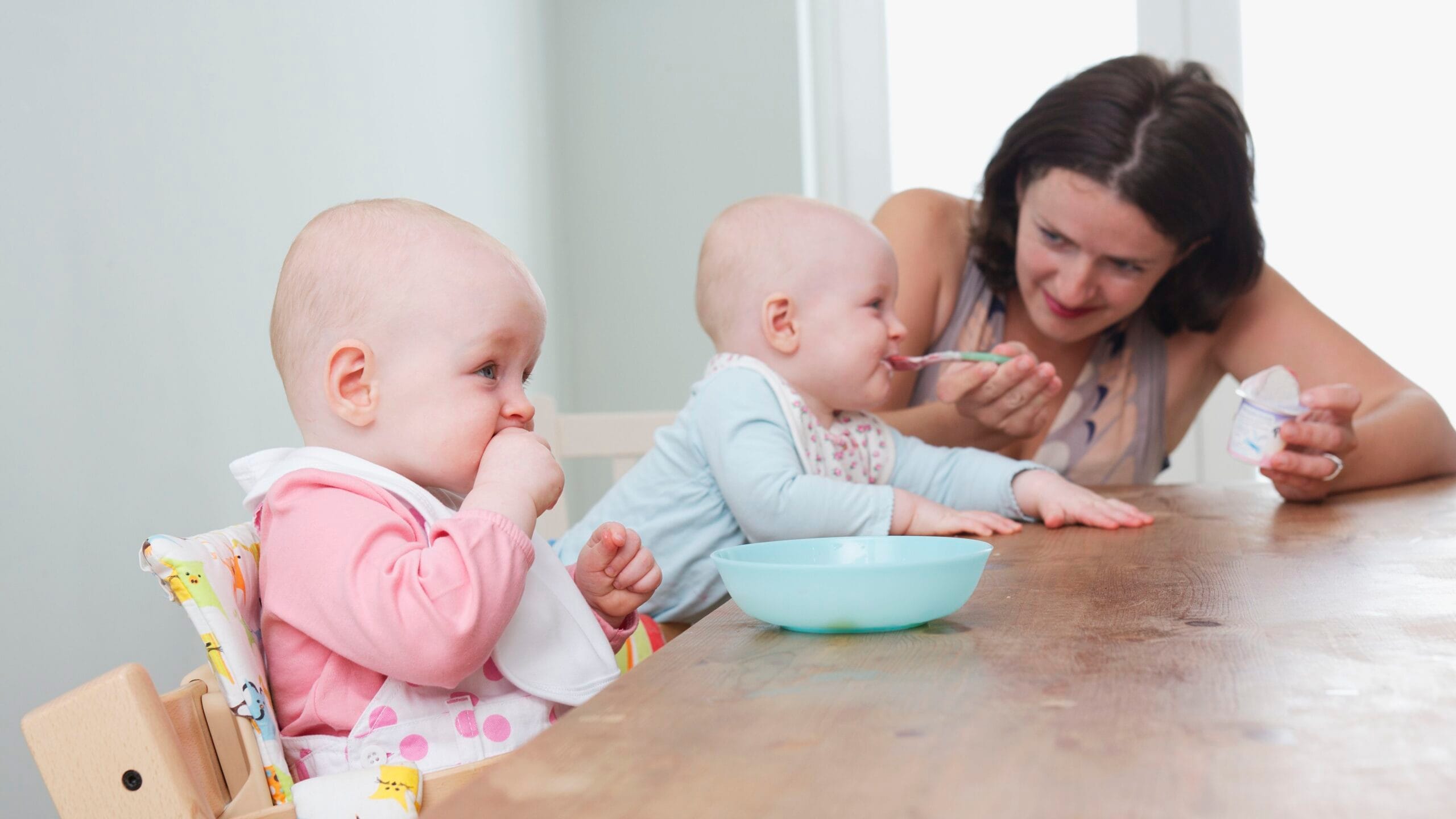 twin baby girls seated at table one being spoon fed the other girl chewing on her fist 104573870 5835b2f83df78c6f6a864398 scaled